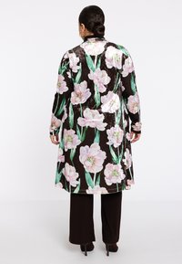 Schwarzer Samt-Langmantel mit pinkfarbigem Pfingstrosen-Blütenmuster, langen Ärmeln und lockerer Passform, kombiniert mit dunkelbraunen Hosen und High Heels.