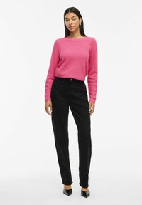 Roze textuurtrui met lange mouwen, gecombineerd met hoge zwarte broek en zwarte puntige hakken, op een effen witte achtergrond.