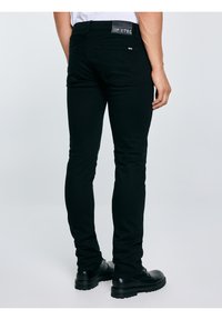 Férfi, aki fekete slim-fit farmert és fekete bőr cipőt visel, hátulról áll egy egyszerű világos háttér előtt.