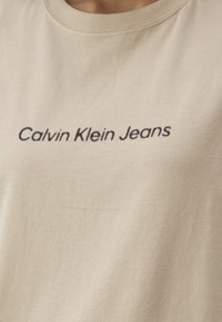 Béžové bavlněné tričko s kulatým výstřihem a krátkými rukávy, na kterém je tmavým písmem natištěno "Calvin Klein Jeans" přes hruď.