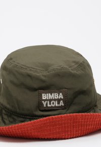 Zielona czapka z rondem w czerwonym prążkowanym stylu, z metką materiałową z napisem "BIMBA Y LOULA". Materiał wydaje się gładki, z widocznymi szczegółami szwów.