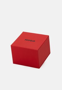 Rote quadratische Box aus Karton mit dem Text "HUGO" in Schwarz auf der obersten Fläche, schräge Sicht, die eine Seite und die offene Klappe zeigt.