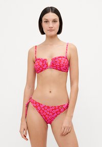 Conjunto de biquíni floral rosa com top franzido, alças ajustáveis e parte de baixo com laços laterais. Apresenta um padrão brilhante e texturizado com detalhes em "SOL".