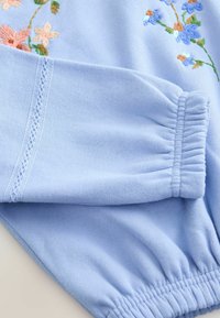 Manchette et poignet en tissu bleu clair avec extrémités élastiques et fleurs brodées roses et bleues sur un côté.