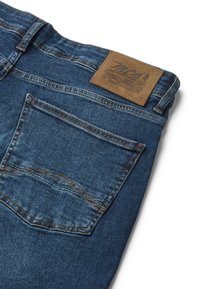 Jean en denim bleu foncé avec une surface texturée. Présente une étiquette en cuir marron à la taille et une poche arrière unique avec des coutures.