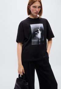 Μαύρο βαμβακερό μπλουζάκι με μεγάλο μονόχρωμο σχέδιο χορευτή μπροστά. Άνετη εφαρμογή με κοντά μανίκια και στρογγυλή λαιμόκοψη.