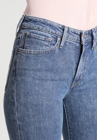 Jeans de mezclilla azul ajustados con cintura alta, diseño de cinco bolsillos, costuras visibles y una ligera decoloración en la textura de la tela.