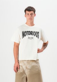 Cream-colored cotton t-shirt with short sleeves, featuring a large black printed "NOTORIOUS" graphic and "WORLD TOUR 1990" detail.