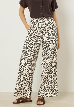 Frau trägt hoch taillierte Hosen mit weitem Bein und weißem Blumen- und Blattmuster auf schwarzem Hintergrund sowie braune Sandalen.