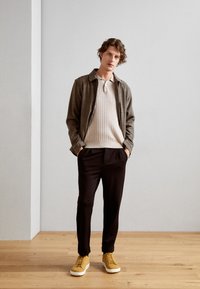 Rutet jacket over en kremfarget kabelstrikket genser, kombinert med svarte bukser og sennepsgule joggesko. Glatt tekstur, skreddersydd passform, moderne design.