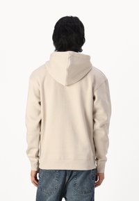 Felpa in cotone beige con cappuccio regolabile, polsini e orlo a coste, caratterizzata da una texture liscia e una vestibilità casual, mostrata da dietro.