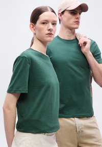 Γυναίκα και άνδρας φορούν ταιριαστά πράσινα μπλουζάκια· ο άνδρας κρατάει ένα μπεζ σακάκι πάνω στον ώμο, φοράει ανοιχτό ροζ καπέλο, και οι δύο κοιτούν μπροστά.