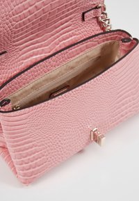 Lyserød tekstureret håndtaske med krokodillemønster, der har en guldtonet lås. Inderummet er beige og afslører rum og en mærkeetiket.