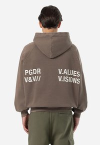 Felpa oversize marrone con cappuccio, caratterizzata da testo bianco sul retro. Le maniche sono in stile raglan e il materiale sembra morbido e strutturato.