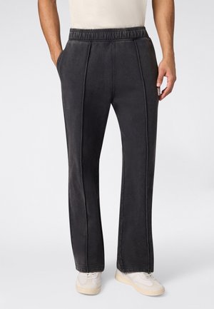 Uomo che indossa pantaloni neri a vestibilità comoda con vita elastica e tasche laterali, abbinati a sneakers bianche, in piedi contro uno sfondo semplice.