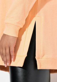 Mano descansando junto a la abertura lateral de una sudadera melocotón, llevada sobre leggings negros brillantes, mostrando textura y contraste de tejidos.