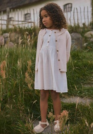 Ein junges Mädchen mit lockigem Haar trägt ein weißes Kleid, eine rosa Strickjacke und Sandalen und steht im hohen Gras nahe einem steinernen Zaun und einem rustikalen Gebäude.