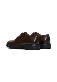Un par de zapatos de vestir de cuero marrón oscuro pulido con suelas negras, mostrados desde la parte trasera y lateral sobre un fondo blanco.