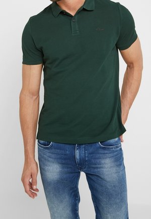 Dunkelgrünes Poloshirt mit Kragen, zwei Knöpfen und einem dezenten Logo. Trägt man mit hellblauen Jeans. Glatte Stofftextur.