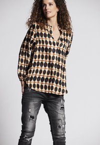 Langärmlige Bluse mit einem geometrischen Muster aus schwarzen und goldenen ovalen Formen auf einem cremefarbenen Hintergrund, kombiniert mit verwaschenen grauen Jeans.