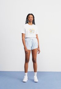 T-shirt en coton blanc avec un graphique floral, associé à un short bleu clair. Baskets blanches et chaussettes de cheville. Coupe simple et décontractée, peu de détails.