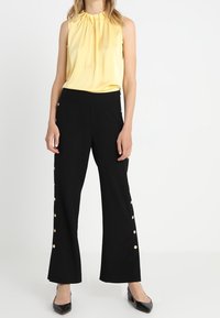 Blusa sin mangas amarilla con escote plisado, combinada con pantalones anchos negros con botones laterales y una textura suave. Bailarinas negras de punta.