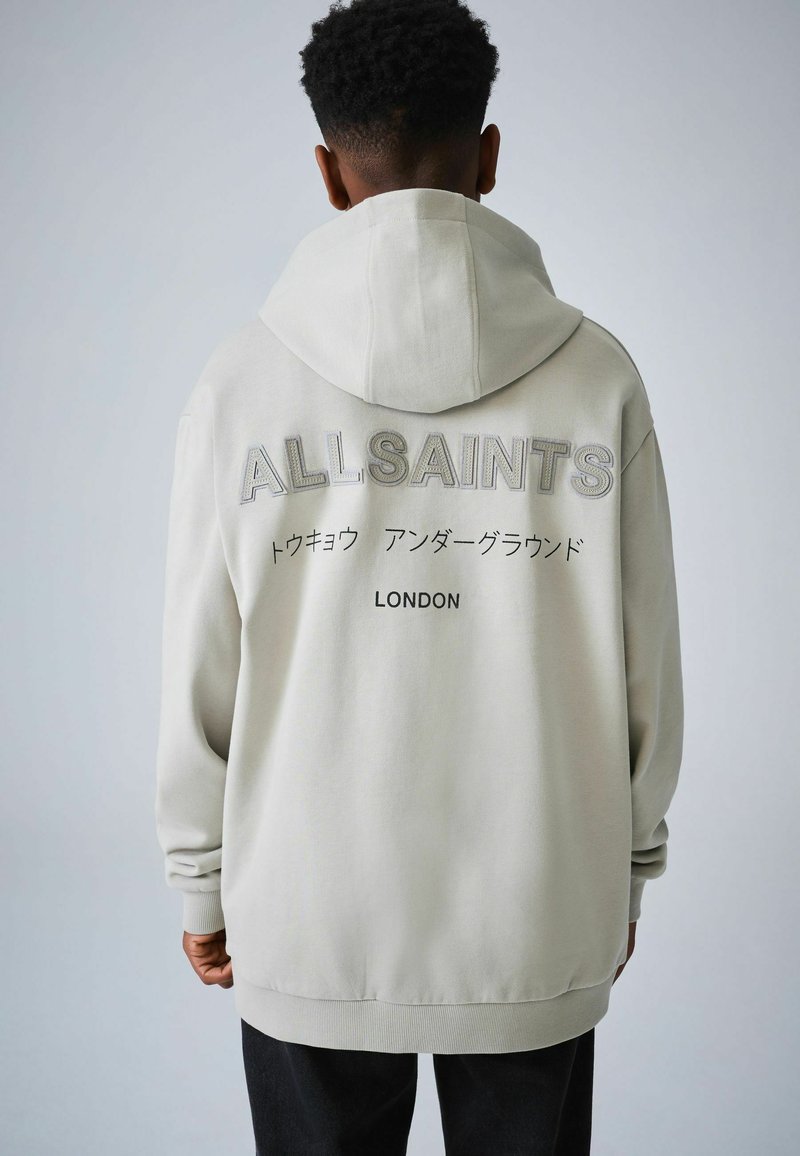 Person mit kurzen lockigen Haaren, die einen hellgrauen Hoodie trägt, auf dessen Rückseite "ALLSAINTS", japanische Schriftzeichen und "LONDON" aufgedruckt sind.