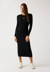 Abito a maglia nero a costine con maniche lunghe, scollo rotondo e vestibilità aderente, con un logo bianco sul petto. Indossato con sneaker bianche.