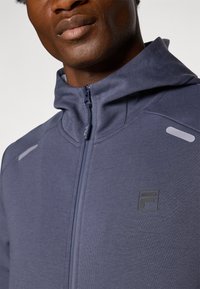 Gros plan d'une personne portant un sweat à capuche bleu avec une fermeture éclair, arborant un petit logo FILA sur la poitrine et des détails réfléchissants sur les épaules.