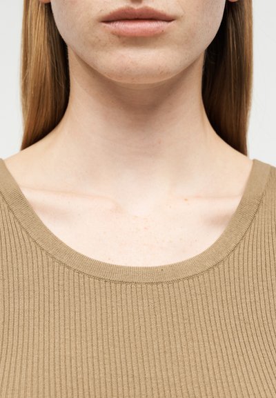 Κοντινό πλάνο του κάτω μέρους του προσώπου, του λαιμού και των ώμων ενός ατόμου που φοράει ένα ριμπ πλεκτό, ανοιχτό καφέ μπλουζάκι με ορατά ίσια μαλλιά.