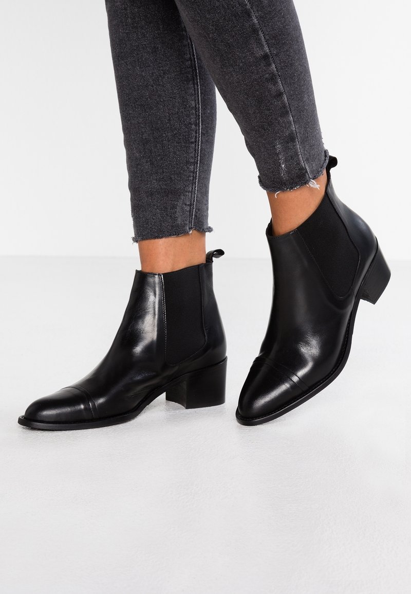 Bottines en cuir noir avec talons carrés bas, portées avec un jean skinny gris foncé à ourlet effiloché.