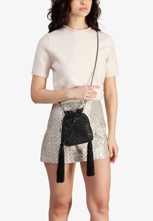 Zwarte pailletten draagkoordtas met twee lange kwastjes, met een glanzende textuur en verstelbare kettingriem, vastgehouden door een persoon in een neutrale outfit.