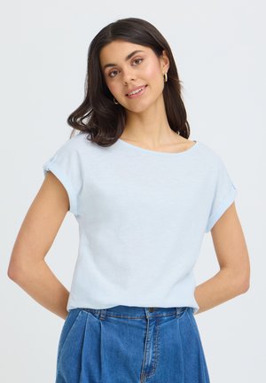Jeune femme aux cheveux foncés souriante, portant un haut bleu clair à manches courtes et un jean bleu taille haute, debout avec les mains derrière le dos.