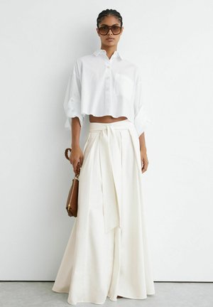 Femme portant un chemisier blanc court et un pantalon blanc taille haute à jambes larges, tenant un sac à main marron, debout contre un mur blanc uni.