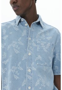 Camicia in denim azzurro chiaro con un motivo a fiori in rilievo, maniche corte, chiusura con bottoni e una tasca sul petto.