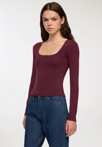 Bordowy top z długim rękawem i kwadratowym dekoltem, z koronkowym wykończeniem i małą kokardką. Wykonany z miękkiego materiału, o dopasowanym kroju. Zestawiony z niebieskimi dżinsami.