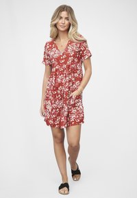 Punainen kukkakuvioinen mekko, jossa on lyhyet hihat, V-aukko, vyötäröä korostava istuvuus ja taskut; kankaassa valkoisia kukkakuvioita; yhdistetty mustiin sandaaleihin.