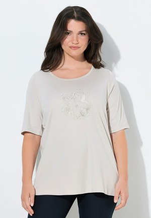 Kvinde med langt brunt hår iført beige kortærmet skjorte med udsmykket blomsterdesign, stående foran en hvid baggrund.