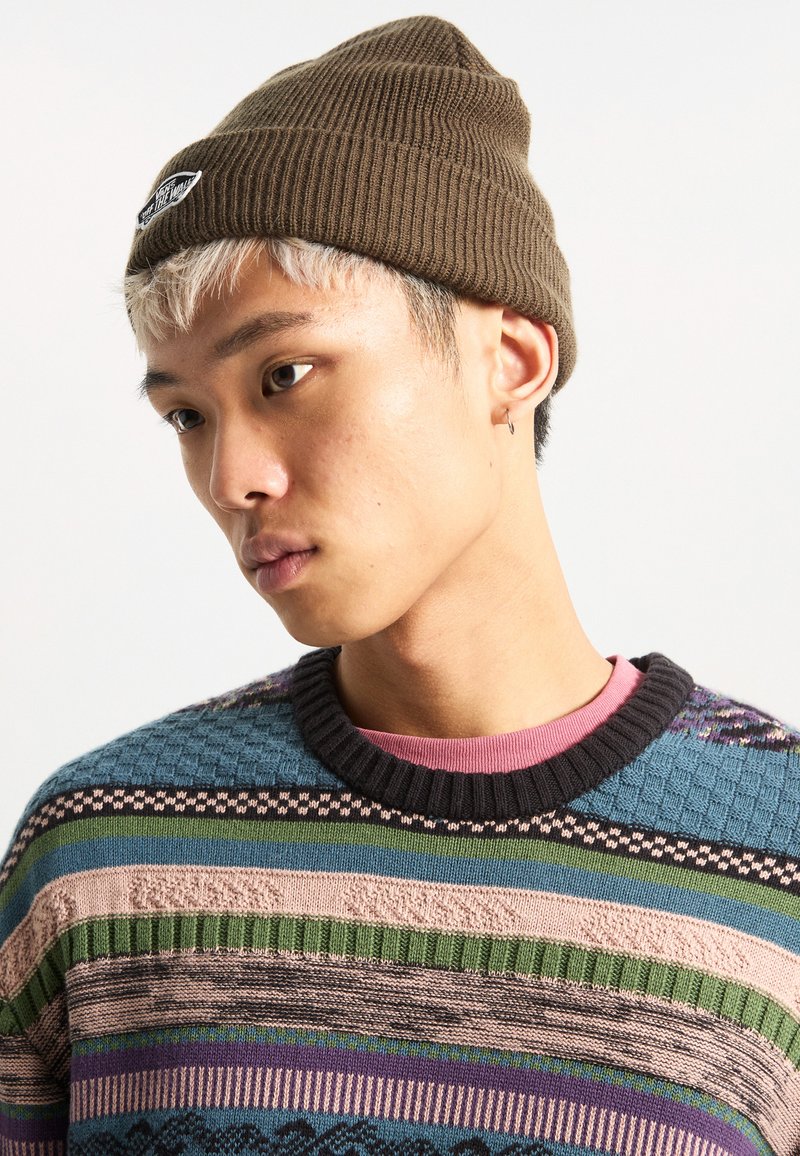 Brown ribbed beanie with logo patch, worn with a multicolored knit sweater featuring various textured patterns and a dark ribbed neckline.