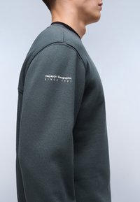 Donkergroen sweatshirt met een ronde hals en geribde boorden. Bevat een logo dat op de linkermouw is gedrukt. Het materiaal lijkt zacht en gestructureerd.