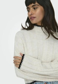 Light beige ribbed sweater with a high collar, featuring black trim on the cuffs and hem. Soft texture and relaxed fit.