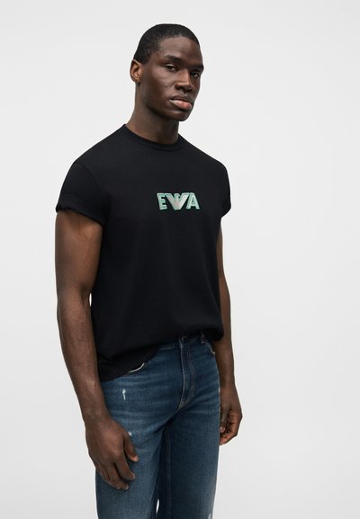 Μαύρο βαμβακερό μπλουζάκι με γυρισμένα μανίκια και πράσινο κεντητό λογότυπο "EVA" στο στήθος, σε συνδυασμό με μπλε τζιν παντελόνι.