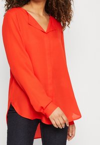Blouse rouge transparente à encolure en V et manches longues. Caractérisée par un ourlet incurvé et des fentes latérales, portée avec un jean foncé.