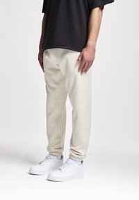 Cremefarbene Jogginghose mit elastischen Bündchen und Taschen, kombiniert mit weißen Sneakern und einem locker sitzenden dunklen Shirt. Glatte Materialoberfläche.