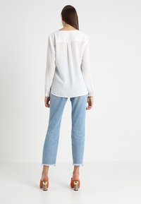 White long-sleeve shirt with a relaxed fit and back yoke detail, paired with light blue frayed hem jeans and orange block-heeled sandals.