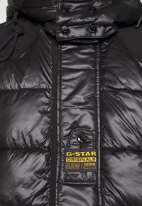 Chaqueta de plumas negra con una textura brillante, que cuenta con un cuello alto, botones a presión y una etiqueta amarilla de marca en la parte frontal.