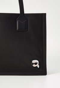 Czarna torba na ramię z skórzanymi uchwytami oraz małą naszywką w kształcie rysunkowego ludka w okularach przeciwsłonecznych i białej chustce w prawym dolnym rogu.