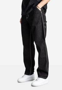 Pantaloni di denim neri con cuciture bianche, taglio dritto, con tasche laterali. Abbinati a sneaker bianche per un contrasto.