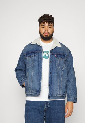 Veste en denim avec un col en fausse fourrure gris clair, fermeture à boutons, deux poches poitrine et une couleur bleu délavé. Associée à un t-shirt blanc.