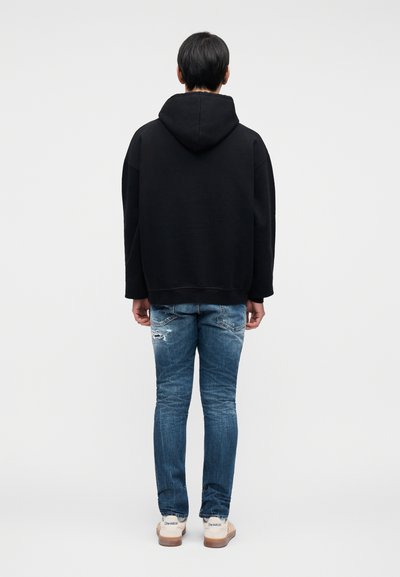 Μαύρο φούτερ με κουκούλα και άνετη εφαρμογή, σε συνδυασμό με φθαρμένο μπλε τζιν και καφέ αθλητικά παπούτσια. Το φούτερ έχει ριμπ τελείωμα και μανσέτες.